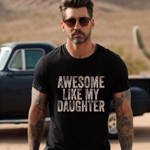 May include: Black crew neck t-shirt with the text "AWESOME LIKE MY DAUGHTER" in a distressed, off-white font. The image shows a person wearing sunglasses and the t-shirt, with a classic car and desert landscape in the background.