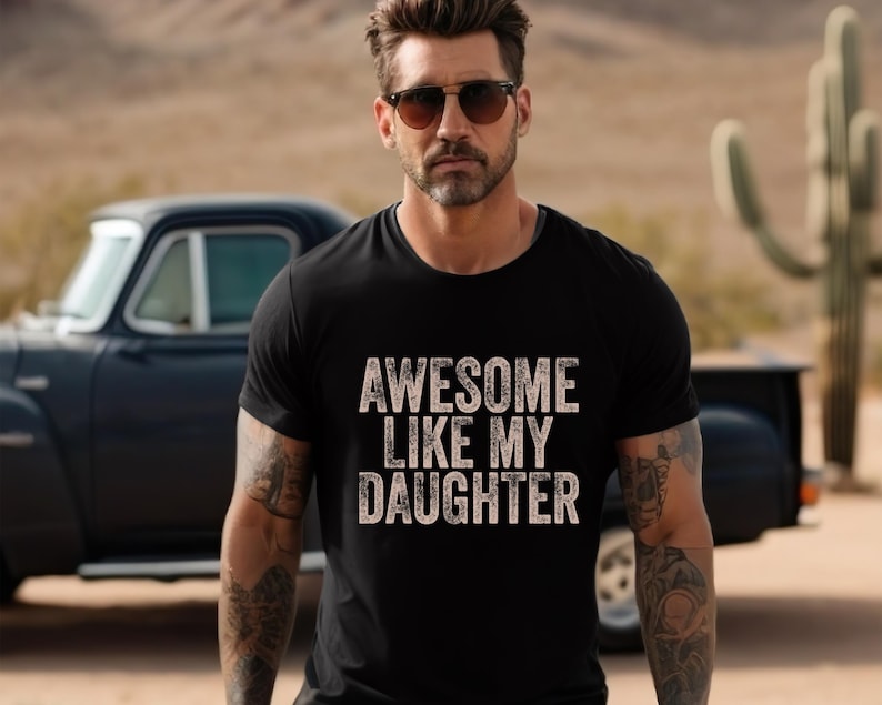 May include: Black crew neck t-shirt with the text "AWESOME LIKE MY DAUGHTER" in a distressed, off-white font. The image shows a person wearing sunglasses and the t-shirt, with a classic car and desert landscape in the background.