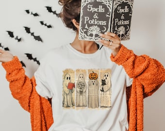 May include: A white t-shirt featuring a graphic of four ghosts over a vintage book page background. One ghost holds a red book, one holds flowers, one holds a jack-o'-lantern, and one holds a black cat. The shirt is worn under an orange cardigan.