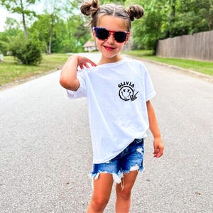 May include: White t-shirt with a black smiley face graphic and the name "Olivia" printed on it. The shirt is being worn by a young person with red and blue sunglasses.