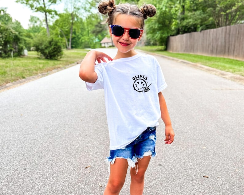 May include: White t-shirt with a black smiley face graphic and the name "Olivia" printed on it. The shirt is being worn by a young person with red and blue sunglasses.