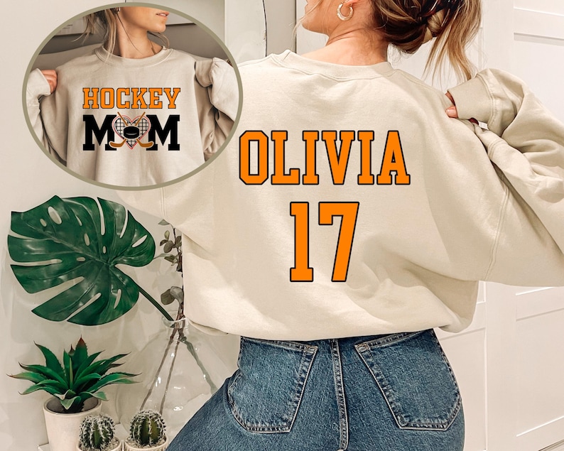May include: A beige sweatshirt with the text "HOCKEY MOM" on the front and "OLIVIA 17" on the back. The sweatshirt has a distressed look and is made of a soft, comfortable fabric.