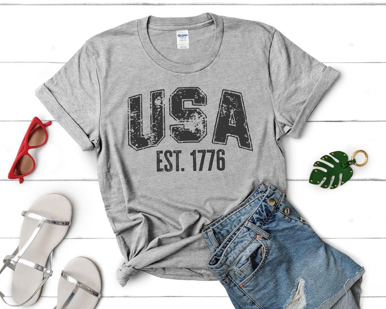 May include: A grey t-shirt with a distressed black graphic that says "USA EST. 1776".