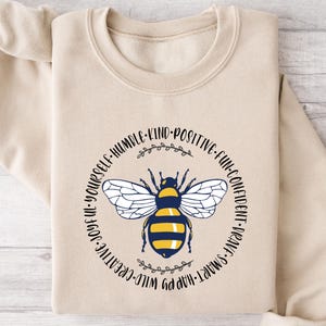 May include: A beige sweatshirt featuring a circular design with a bee illustration. The bee is black, yellow, and white, surrounded by text that reads "Be Yourself, Humble, Kind, Positive, Fun, Confident, Brave, Smart, Happy, Wild, Creative, Joyful."