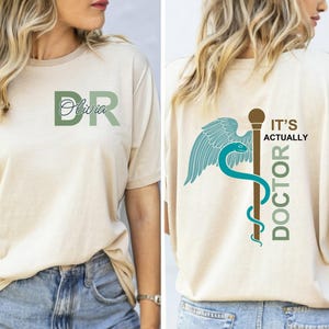 May include: A beige short-sleeve t-shirt featuring a green DR monogram with the name Olivia on the front. The back displays a medical caduceus symbol with wings and a snake, accompanied by the text IT'S ACTUALLY DOCTOR in green and brown.