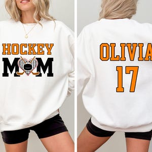 Puede incluir: Sudadera blanca con el texto "HOCKEY MOM" en negro y naranja con un gr&aacute;fico de un palo de hockey y un disco. La parte posterior de la sudadera tiene el texto "OLIVIA 17" en naranja.