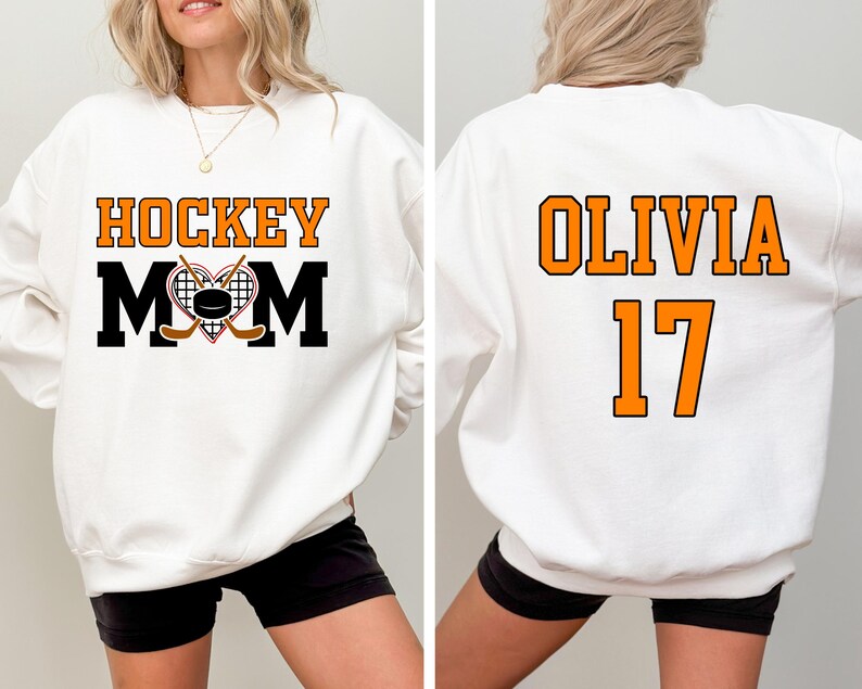 Puede incluir: Sudadera blanca con el texto "HOCKEY MOM" en negro y naranja con un gr&aacute;fico de un palo de hockey y un disco. La parte posterior de la sudadera tiene el texto "OLIVIA 17" en naranja.