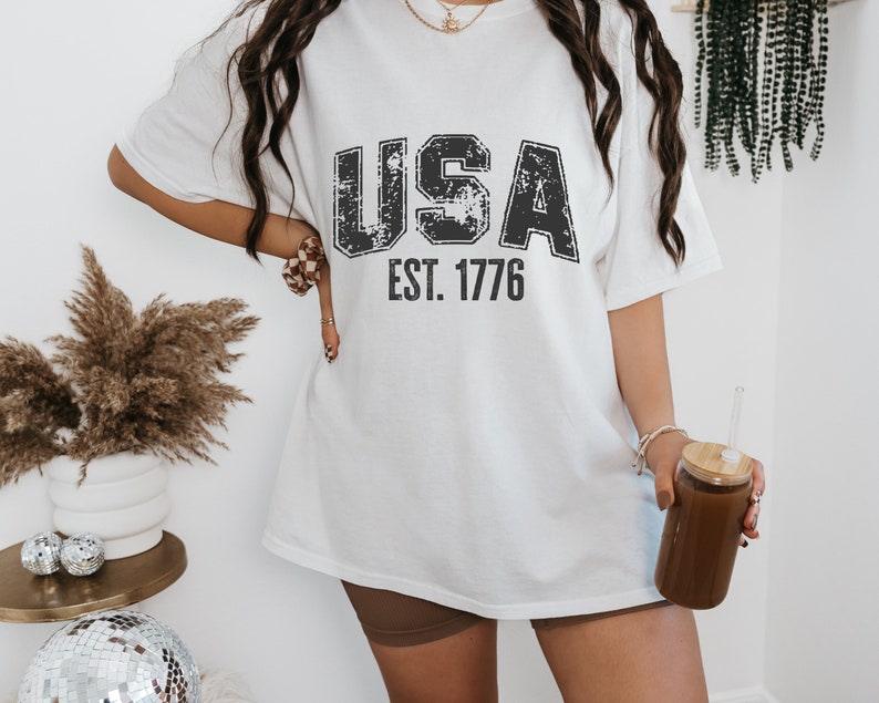 May include: White oversized t-shirt with a distressed black graphic that says "USA EST. 1776".