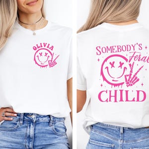 May include: White t-shirt with a pink smiley face with crossed eyes and a skeleton hand making a peace sign. The text "Olivia" is above the smiley face. The back of the shirt has a pink smiley face with crossed eyes and a skeleton hand making a peace sign. The text "Somebody's Feral Child" is above the smiley face.