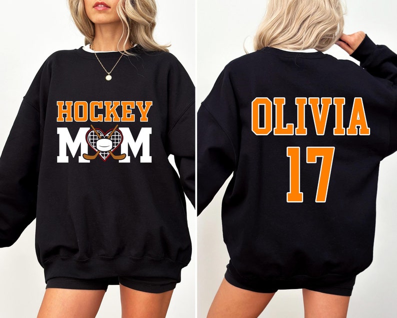 Puede incluir: Sudadera negra con el texto "HOCKEY MOM" en blanco con un coraz&oacute;n y palos de hockey en naranja. La parte posterior de la sudadera tiene el texto "OLIVIA 17" en naranja.