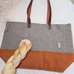 May include: A gray and brown tote bag with leather handles. The bag has a brown fabric bottom and a small brown tag with the text "Daily Grind" on it. A skein of yarn in shades of yellow, orange, and pink is in front of the bag.