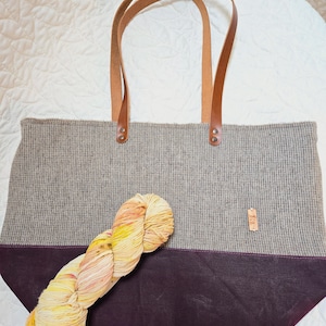 May include: A gray and purple tote bag with leather straps and a skein of yellow, orange, and pink yarn. The bag has a small brown label with text that is too small to read.