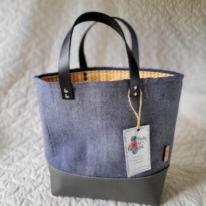 May include: A blue and gray tote bag with a yellow and white patterned lining. The bag has black leather handles and a leather bottom. A tag with the text "Strung Out Knits" is attached to the bag.