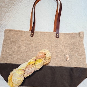 May include: A beige tote bag with brown leather handles and a brown canvas bottom. The bag is filled with a skein of yellow, orange, and pink yarn.