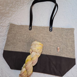 May include: A beige tote bag with black leather handles and a dark brown bottom. The bag is filled with a skein of yellow and orange yarn.