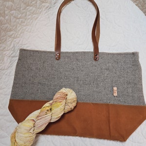 May include: A gray and brown tote bag with leather handles. The bag has a brown canvas bottom and a gray tweed top. A skein of yellow, pink, and white yarn is sitting on the bag.