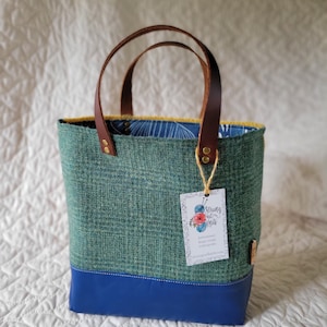 May include: A teal green tweed tote bag with a blue leather bottom and brown leather handles. The bag has a yellow and white floral lining and a small tag that reads "Strong Knit Vatis" with a website address.