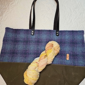 May include: A large tote bag with a blue and purple plaid pattern, a dark green bottom, and black leather handles. A skein of colorful yarn rests on the bag. A small tag is attached to the bag.