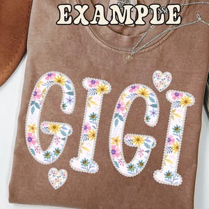 May include: A brown t-shirt with the word "GIGI" in large, floral-patterned letters. The letters are white with a floral design of yellow, pink, and blue flowers. Two small heart shapes with the same floral pattern are also present.