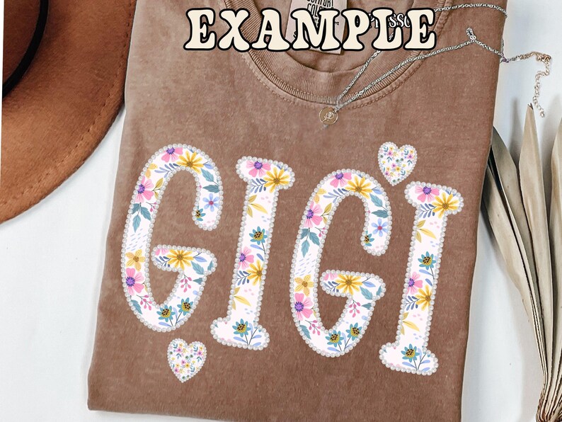 May include: A brown t-shirt with the word "GIGI" in large, floral-patterned letters. The letters are white with a floral design of yellow, pink, and blue flowers. Two small heart shapes with the same floral pattern are also present.
