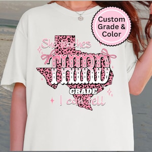 May include: White t-shirt featuring a pink and black leopard print Texas outline with the words "She teaches third grade, I can tell." The design includes pink bows and stars. A pink circle reads "Custom Grade & Color."