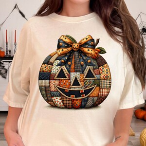 Puede incluir: Camiseta color crema con un diseño de calabaza de retazos. La calabaza tiene una cara sonriente de Jack-o'-lantern y un lazo decorativo. El diseño incorpora varios patrones y colores, perfecto para Halloween.