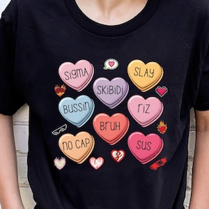 Może przedstawiać: Czarny t-shirt z kolorowymi cukierkami w kształcie serca z slangowymi terminami takimi jak "SIGMA" i "SLAY". Projekt zawiera małe elementy graficzne, takie jak płomienie i serca, tworząc zabawną i modną estetykę.