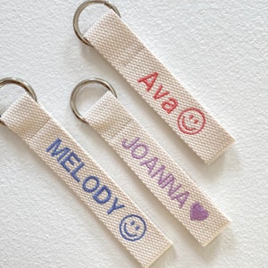 May include: Three white canvas keychains with metal rings. The keychains have different names embroidered on them in different colors: "Ava" in pink with a smiley face, "Joanna" in purple with a heart, and "Melody" in blue with a smiley face.