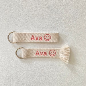 May include: Two white canvas keychains with a silver ring. The keychains have the name "Ava" and a red smiley face embroidered on them. One keychain has a white tassel.