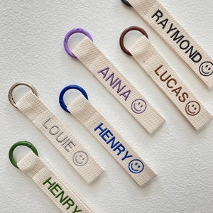 May include: Six keychains with different colored rings and embroidered names: Raymond, Lucas, Anna, Henry, Louie, and Henry. Each keychain has a smiling face embroidered on the fabric.