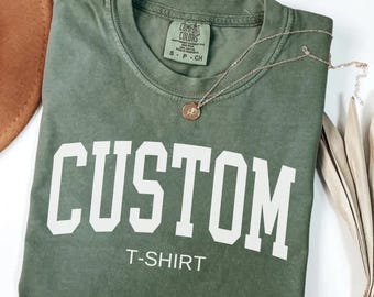 May include: A moss green t-shirt with the word "CUSTOM" printed in white on the front. The text "T-SHIRT" is printed below the word "CUSTOM".