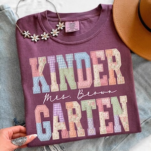 May include: A maroon-colored Comfort Colors t-shirt with the words "KINDER GARDEN" in patchwork letters. The shirt also features the name "Mrs. Brown" in script. A floral necklace and a brown hat are also visible.