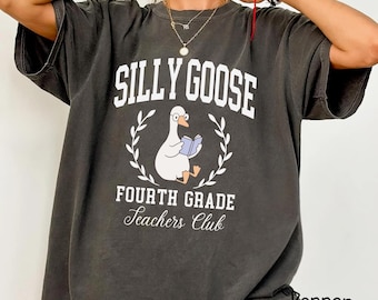 Puede incluir: Camiseta gris oscuro con las palabras "SILLY GOOSE" en blanco sobre un ganso de dibujos animados leyendo un libro. Debajo, "FOURTH GRADE Teachers Club" en escritura blanca. La camiseta es de corte holgado.