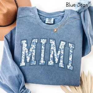 May include: Blue jean sweatshirt with a white floral print that says "MIMI" in a collegiate style font.