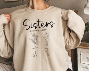 May include: A sand-colored sweatshirt with the text "Sisters blooming together" and two stylized flowers with the names "Emily" and "Michaela" written underneath.