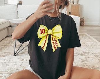 May include: Black t-shirt featuring a large yellow bow design with red baseball stitching. The bow has the number "10" and the name "Jessica" printed on it. The shirt is a casual style.
