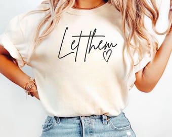May include: A white t-shirt with the text "Let them" in black script with a small heart symbol. The shirt is being worn by a person with long blonde hair. The text "Ivory" is visible in the bottom right corner of the image.