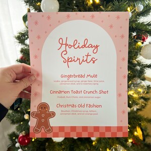 Op de afbeelding: Een roze en witte printbare kaart met de tekst "Holiday Spirits" in rood. De kaart vermeldt drie feestelijke cocktailrecepten: Gingerbread Mule, Cinnamon Toast Crunch Shot en Christmas Old Fashion.