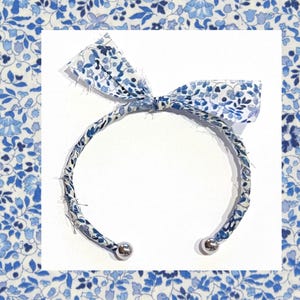 Puede incluir: Una diadema floral azul con un lazo. La diadema está hecha de tela con un estampado floral blanco y azul. Los extremos de la diadema tienen detalles de bolas de metal plateado.