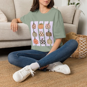 May include: A sage green short-sleeve t-shirt featuring a graphic print of nine pumpkins in various patterns, including floral, plaid, leopard print, and cow print, arranged over three vertical light purple stripes with white polka dots.
