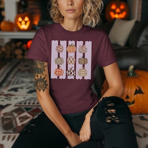 May include: A maroon short-sleeve t-shirt featuring a graphic print of nine pumpkins arranged in three vertical columns. The pumpkins display various patterns including floral, plaid, leopard print, and cow print, set against a light purple background.