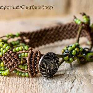 May include: A handmade macrame bracelet featuring olive green beads and a brown woven design. The bracelet has a decorative silver button clasp and is accented with additional green beads. The bracelet is displayed on a wooden surface.