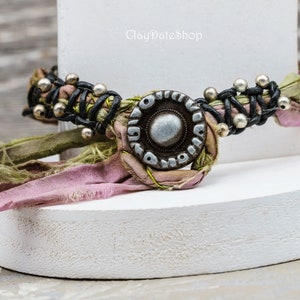 May include: A pink and green fabric bracelet with a silver and black metal charm. The bracelet is tied with a black leather cord and has silver beads.