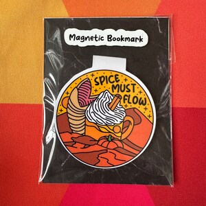 May include: A magnetic bookmark featuring a cartoon illustration of a pumpkin spice latte with the text "Spice Must Flow". The bookmark is on a black background with the text "Magnetic Bookmark" at the top.