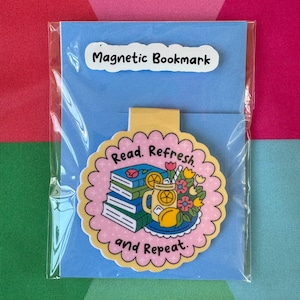 May include: A magnetic bookmark with a scalloped edge, featuring a stack of books, a lemonade drink, and flowers. The text "Read. Refresh. and Repeat." is displayed. The bookmark is packaged on a blue card with the text "Magnetic Bookmark".