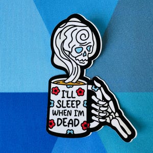 May include: A white coffee mug with a skeleton holding it. The mug has red and blue flowers and the text "I'll sleep when I'm dead".