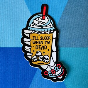 May include: A yellow and blue cartoon illustration of a skeleton hand holding a coffee cup with whipped cream and the text "I'll sleep when I'm dead".
