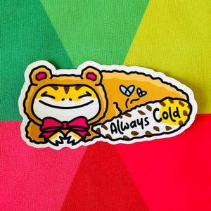 May include: A sticker featuring a cartoon frog wearing a tiger costume and a pink bow tie. The frog is smiling, and a speech bubble says "Always Cold". The sticker has a yellow, brown, and white color scheme.
