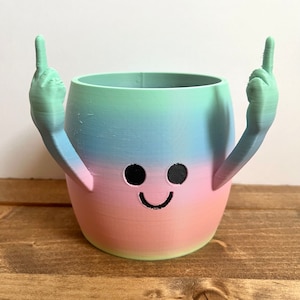 May include: A colorful ombre planter with a smiley face and two raised middle fingers. The planter is made of ceramic and has a smooth, glossy finish.