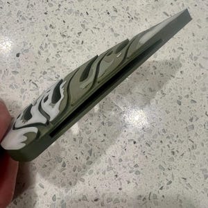 Puede incluir: Una funda de tel&eacute;fono de camuflaje verde y blanco con una ranura negra en el lateral.